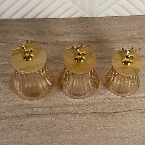 Three small ribbed glass jars with light bamboo lids and gold reindeer finials, arranged on a wooden surface against a white paneled background.