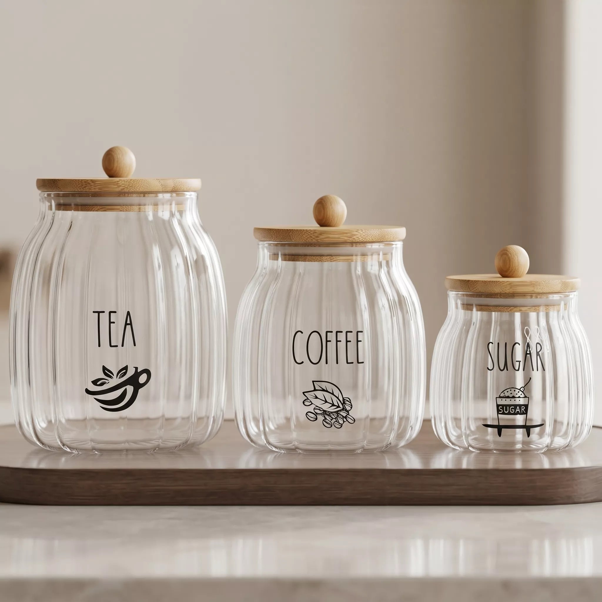 Three ribbed clear glass canisters with light wood lids and round wooden knobs, labeled TEA, COFFEE, and SUGAR in black text, arranged on a wooden tray against a soft neutral background.