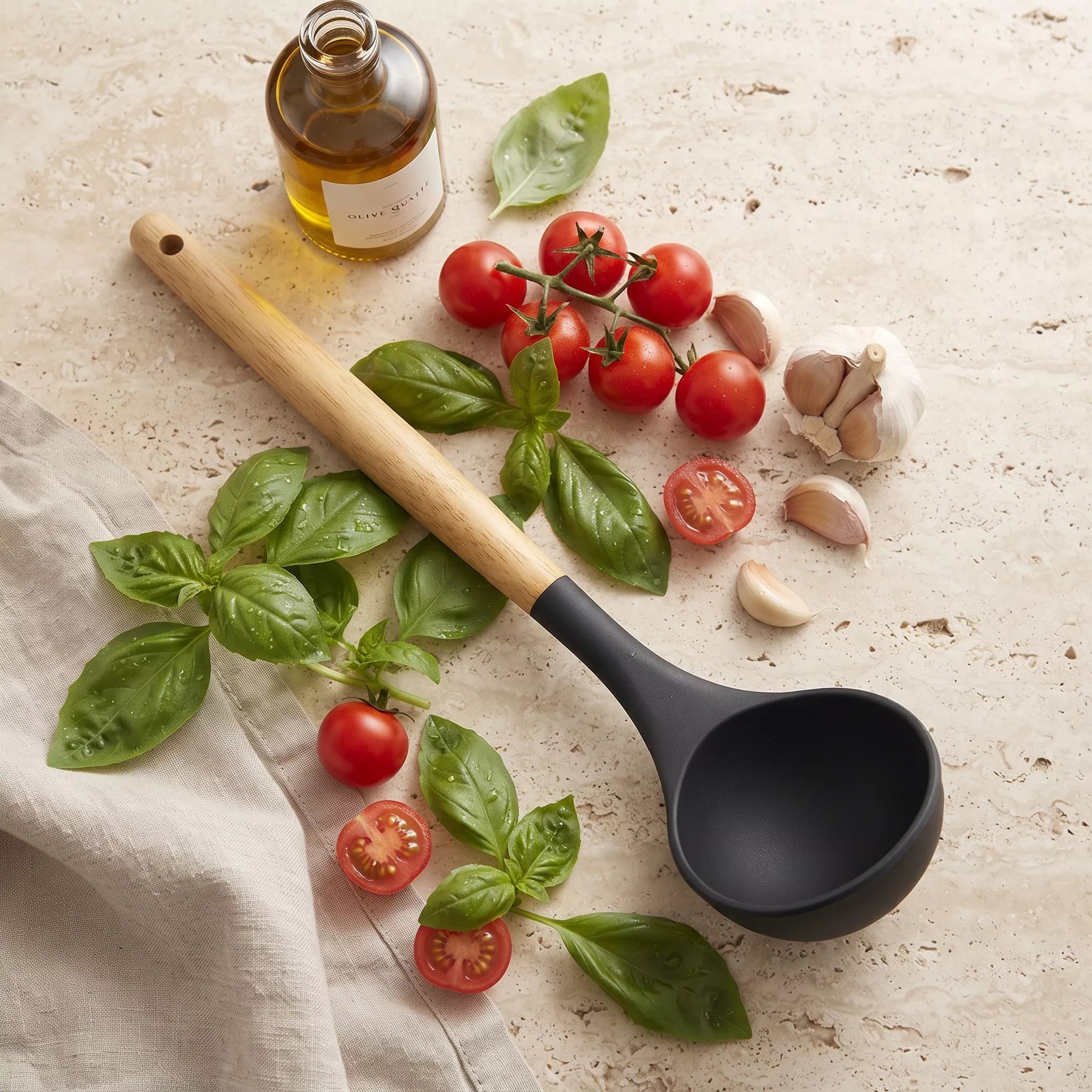 A black silicone ladle with a light wood handle is arranged on a light stone surface with fresh basil leaves, cherry tomatoes, garlic, and a bottle of oil as styled props.