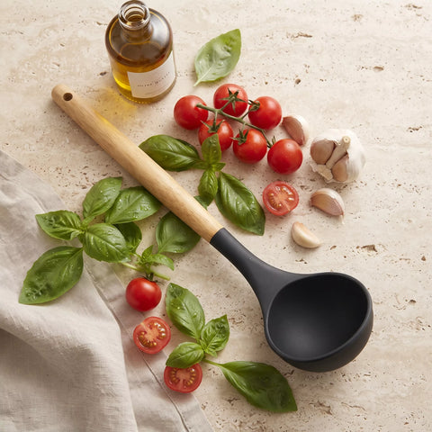 A black silicone ladle with a light wood handle is arranged on a light stone surface with fresh basil leaves, cherry tomatoes, garlic, and a bottle of oil as styled props.