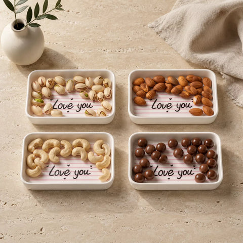 Four small rectangular white ceramic trays arranged in a 2x2 grid on a beige stone-like surface. Each tray contains different snacks: pistachios, almonds, cashews, and chocolate candies.