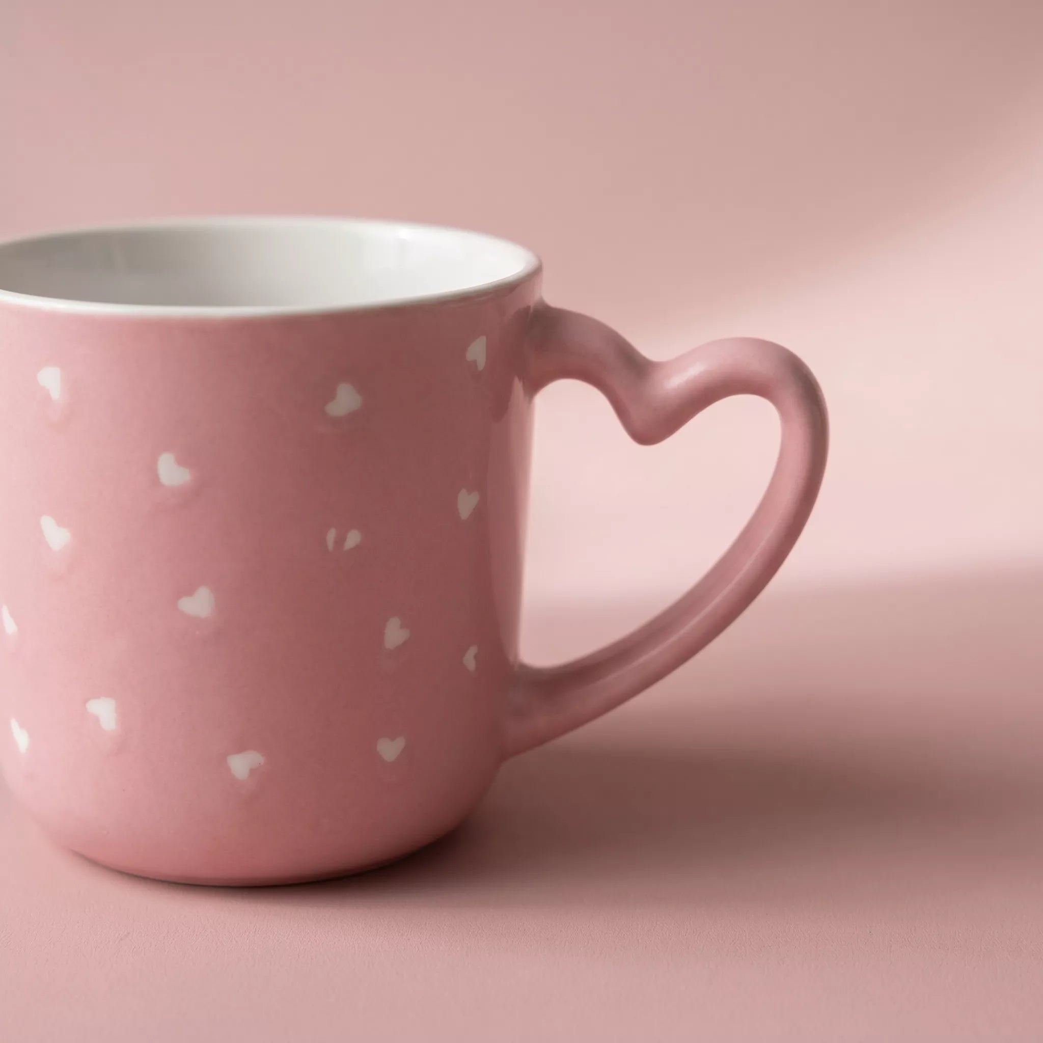 Pink mug with heart-shaped handle and white heart patterns on a pink background