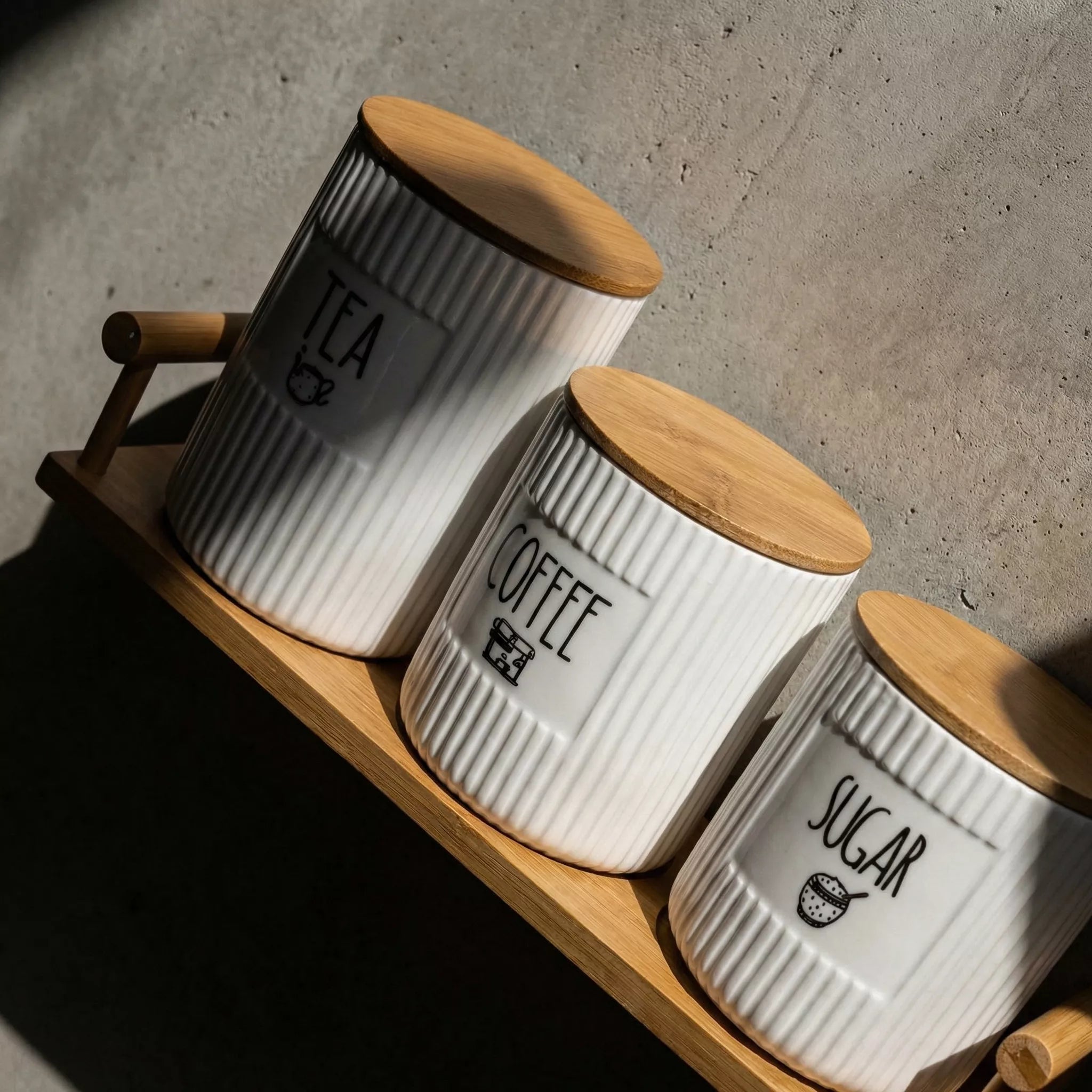 Three white canisters with wooden lids labeled 'Tea', 'Coffee', and 'Sugar' on a wooden tray.