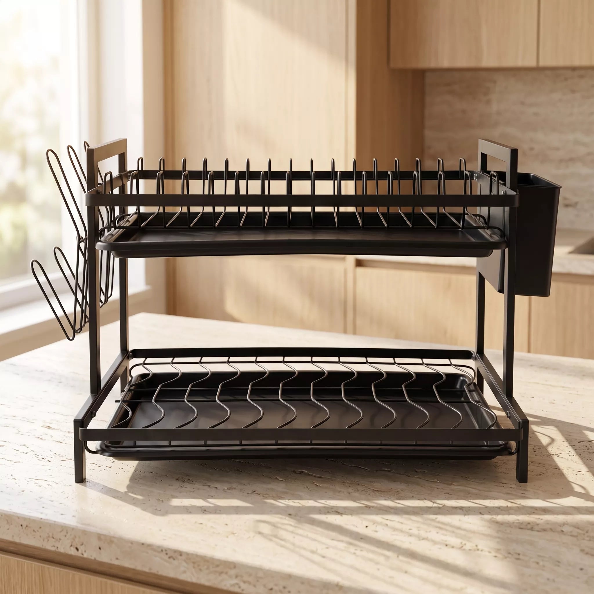 A black metal two-tier dish rack with a side cup and utensil holder, shown on a kitchen countertop in a warm, sunlit kitchen setting.