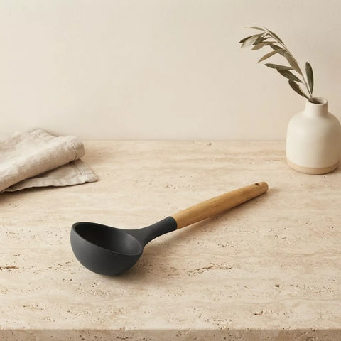 A matte black ladle with a light natural wood handle resting on a textured beige stone surface, with a folded linen cloth and a small vase with dried leaves in the background.