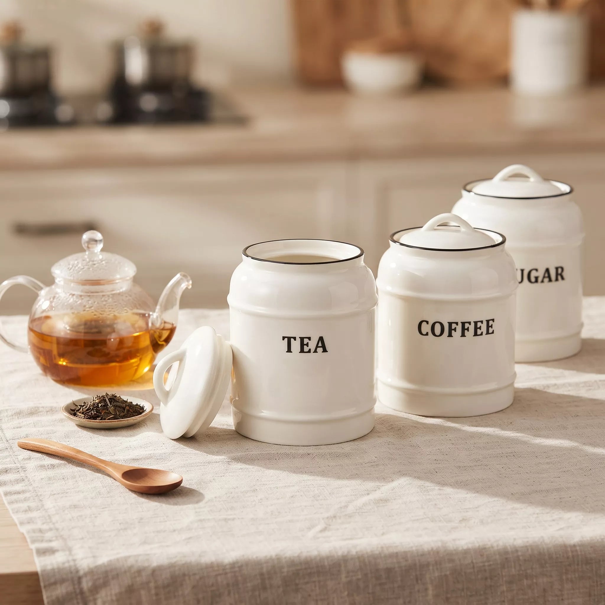 Three white enamel canisters labeled TEA, COFFEE, and SUGAR sit on a beige tablecloth beside a glass teapot and wooden spoon in a softly blurred kitchen setting.