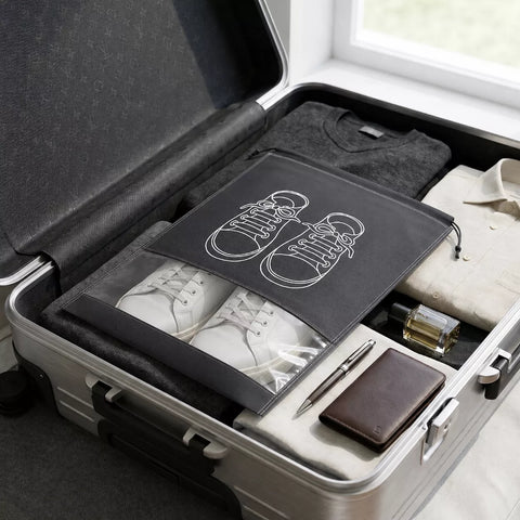 A gray fabric shoe bag with white line-art shoe illustrations is shown inside an open silver suitcase, alongside folded clothing, white sneakers, a pen, a wallet, and a small bottle.