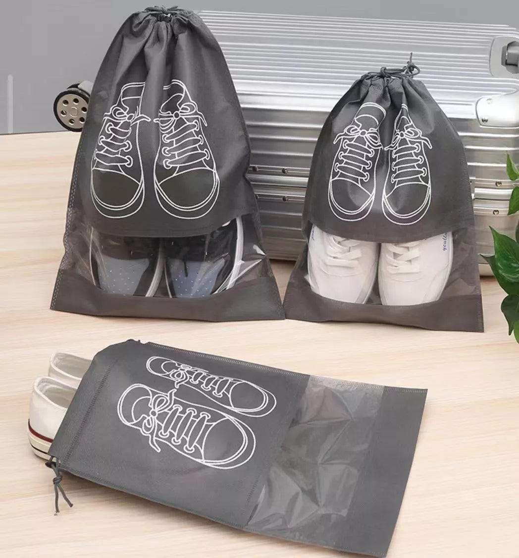 Three dark gray shoe storage bags with clear side windows and white shoe-illustration prints, shown with shoes inside on a light wood surface in front of a corrugated metal panel.
