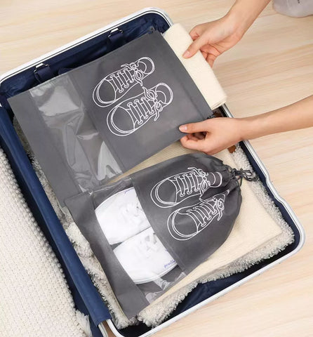 A person's hands are shown packing travel shoe bags into an open suitcase. The bags are dark gray with white shoe line drawings and include one transparent shoe bag holding a pair of white sneakers and one drawstring shoe bag.