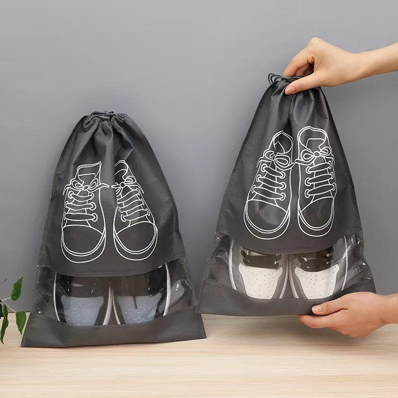 Two black drawstring shoe bags with clear plastic lower sections and white shoe line drawings, shown on a light wood surface against a gray wall. One bag is standing on the floor and the other is being held up by hands to demonstrate its stretch.