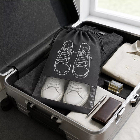 A black drawstring shoe bag with a white sneaker outline graphic is shown inside an open silver suitcase, alongside white sneakers, a folded cream shirt, a perfume bottle, a pen, and a brown wallet.