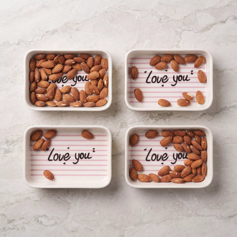 Four small square white dishes arranged in a 2x2 grid on a light marble surface. Each dish contains almonds and has the phrase 'Love you' written in black script over a pale pink lined pattern.