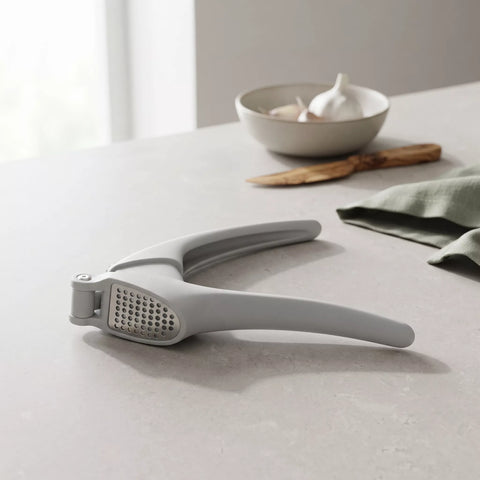 A light gray garlic press resting on a light gray countertop, with a blurred bowl of garlic and a wooden spoon in the background.