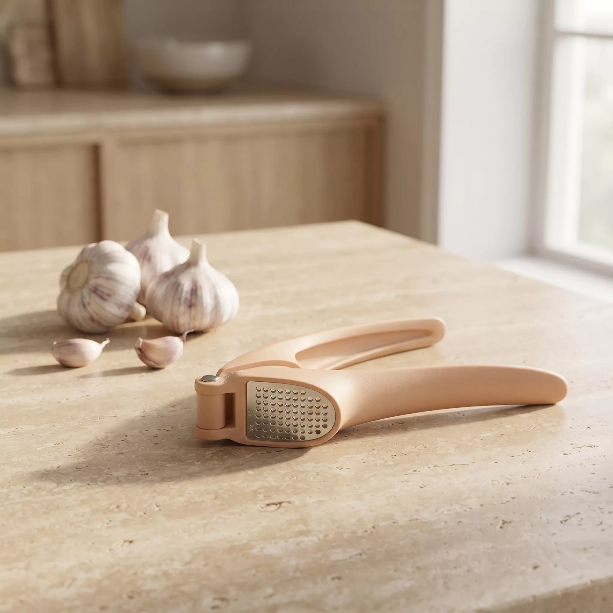 A beige garlic press and grater tool sits on a light stone countertop with garlic bulbs and cloves nearby, in a softly lit kitchen setting.