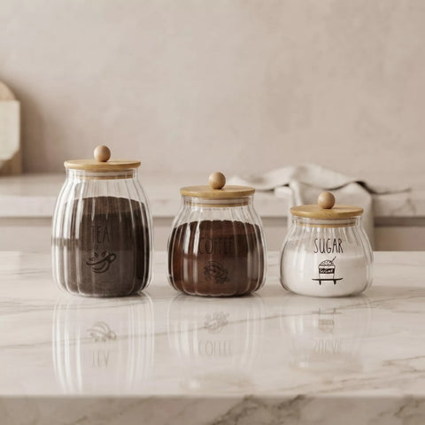 Three ribbed clear glass canisters with light wood lids and wooden knobs, labeled TEA, COFFEE, and SUGAR, arranged on a white marble countertop against a warm beige background.