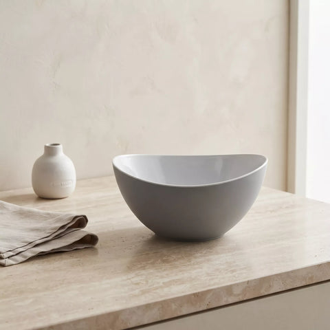 A matte gray bowl with a wide, rounded shape sits on a light beige stone countertop. A folded beige cloth and a small white ceramic bottle are placed beside it against a softly textured beige wall.