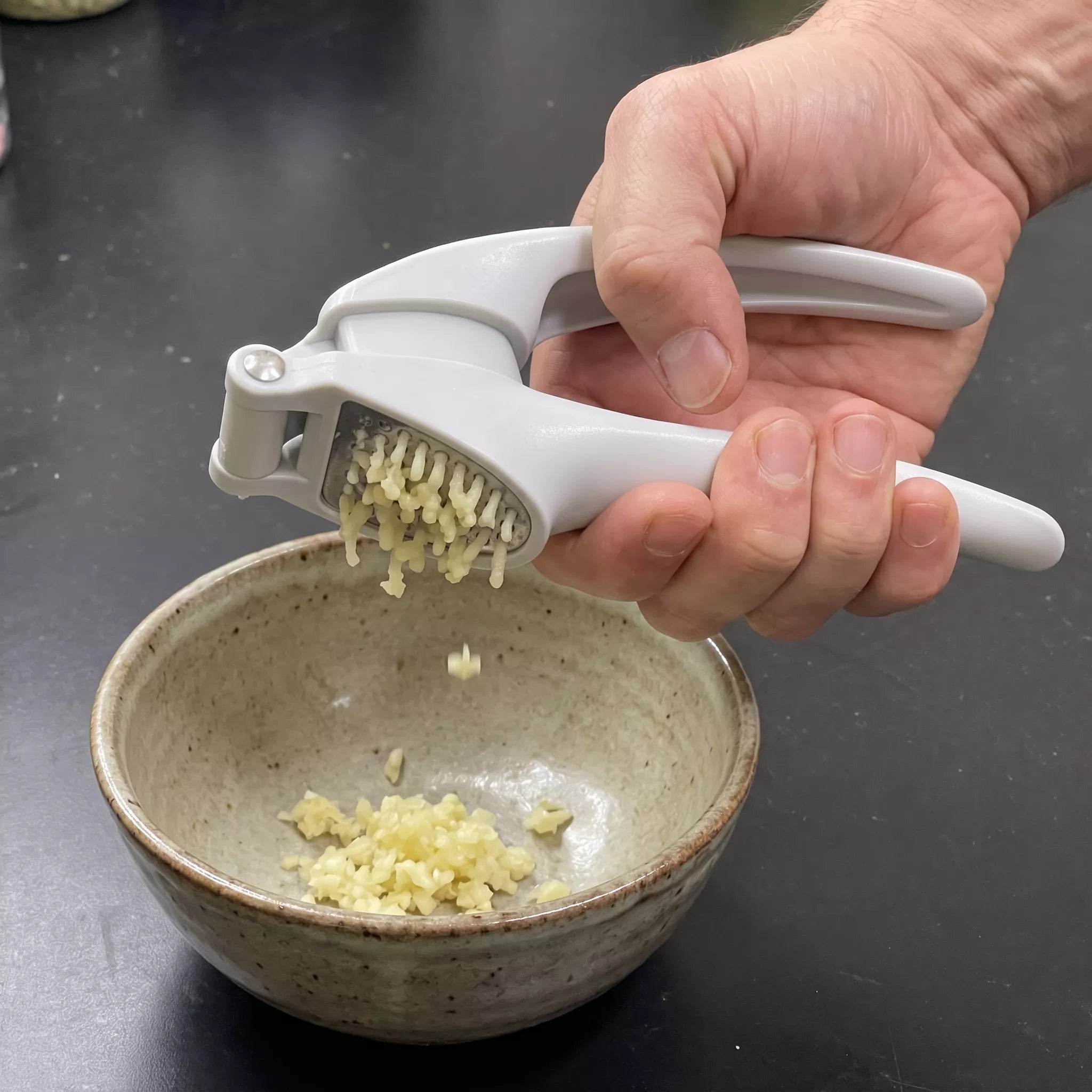 A hand using a white garlic press to mince garlic over a speckled beige ceramic bowl on a dark countertop.