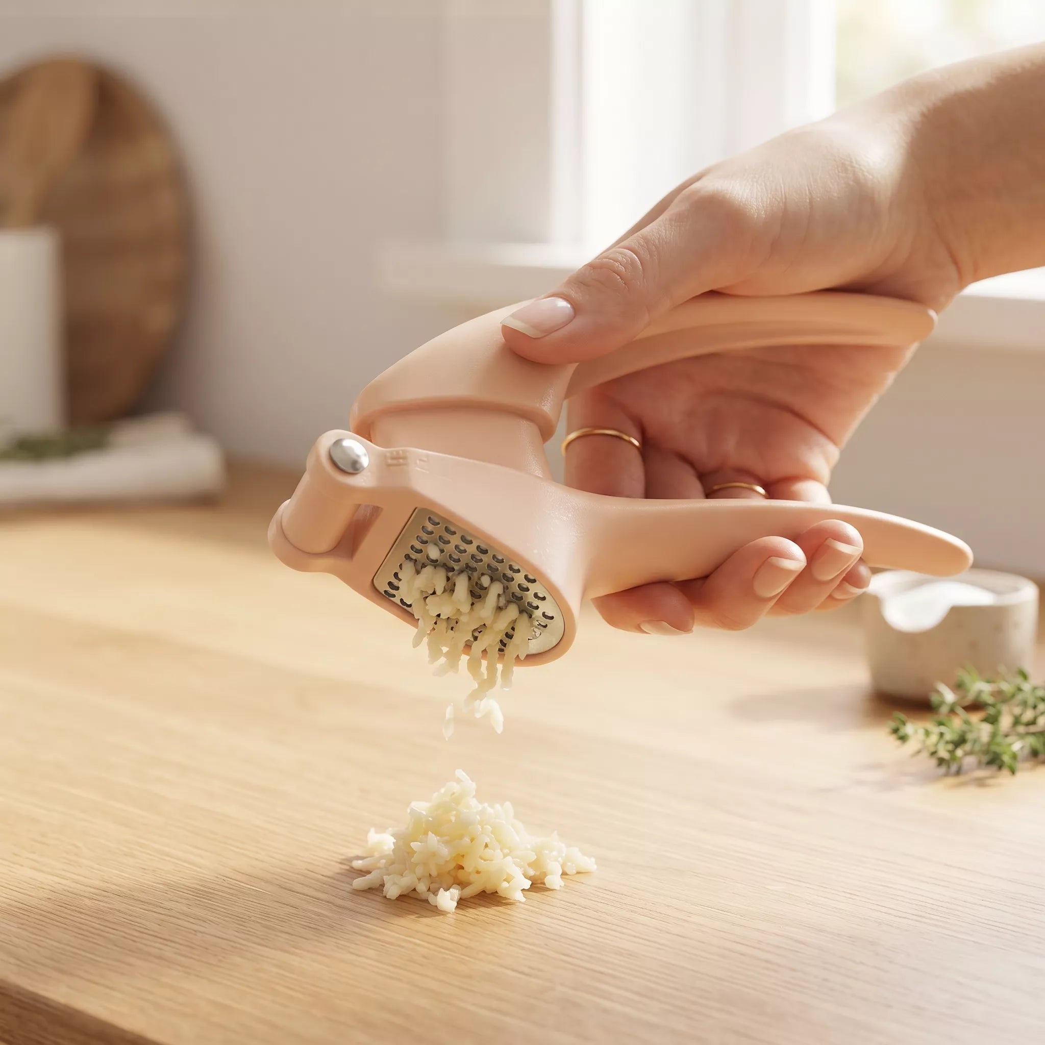 A hand using a peach-colored garlic press to crush garlic on a wooden countertop, with minced garlic falling into a pile below.