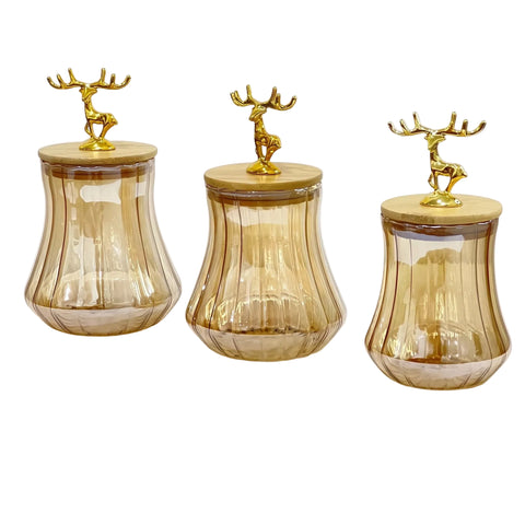 Three amber glass storage jars with fluted sides and light wood lids, each topped with a small gold deer figurine, shown on a white background.