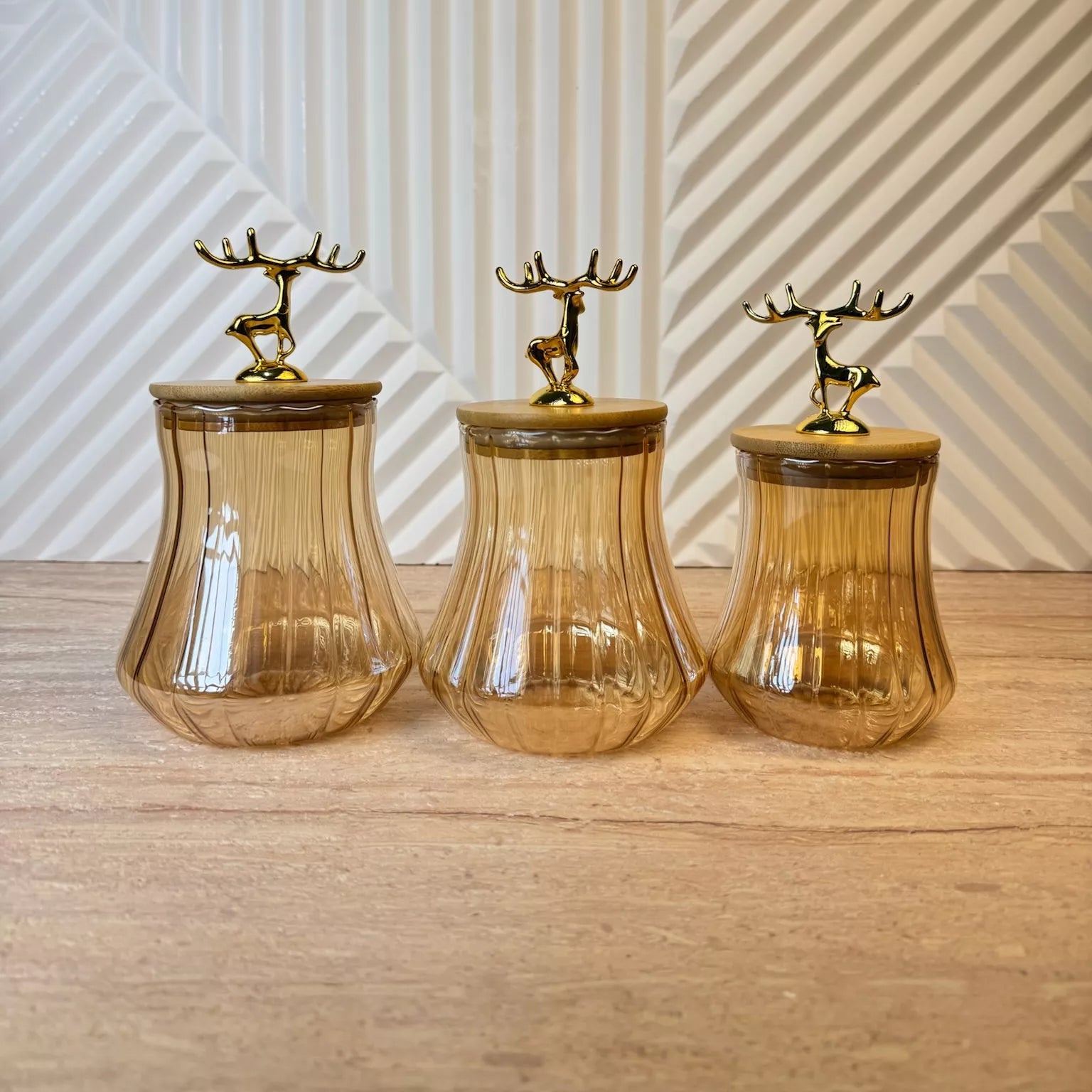 Three amber glass canisters with ribbed flared bases and lids topped by gold deer figurines, arranged on a light wood surface against a geometric patterned wall.