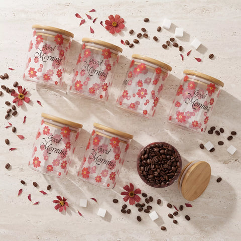 Six clear glass canisters with light wood lids are arranged on a light stone surface with scattered coffee beans, flower petals, and sugar cubes. The canisters are printed with red and pink cherry blossom flowers and the words 'Good Morning' in black script.