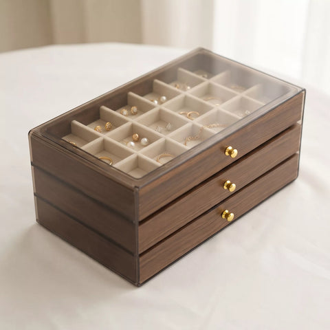 A rectangular jewelry organizer box with a dark brown wood-grain exterior, three drawers with gold-tone knobs, and a clear glass lid showing multiple cream-colored compartments filled with earrings and jewelry.