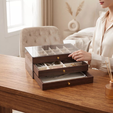 A woman in a cream satin shirt organizes jewelry in a dark wood drawer organizer with a clear top lid and multiple compartments, set on a wooden table in a softly lit bedroom.