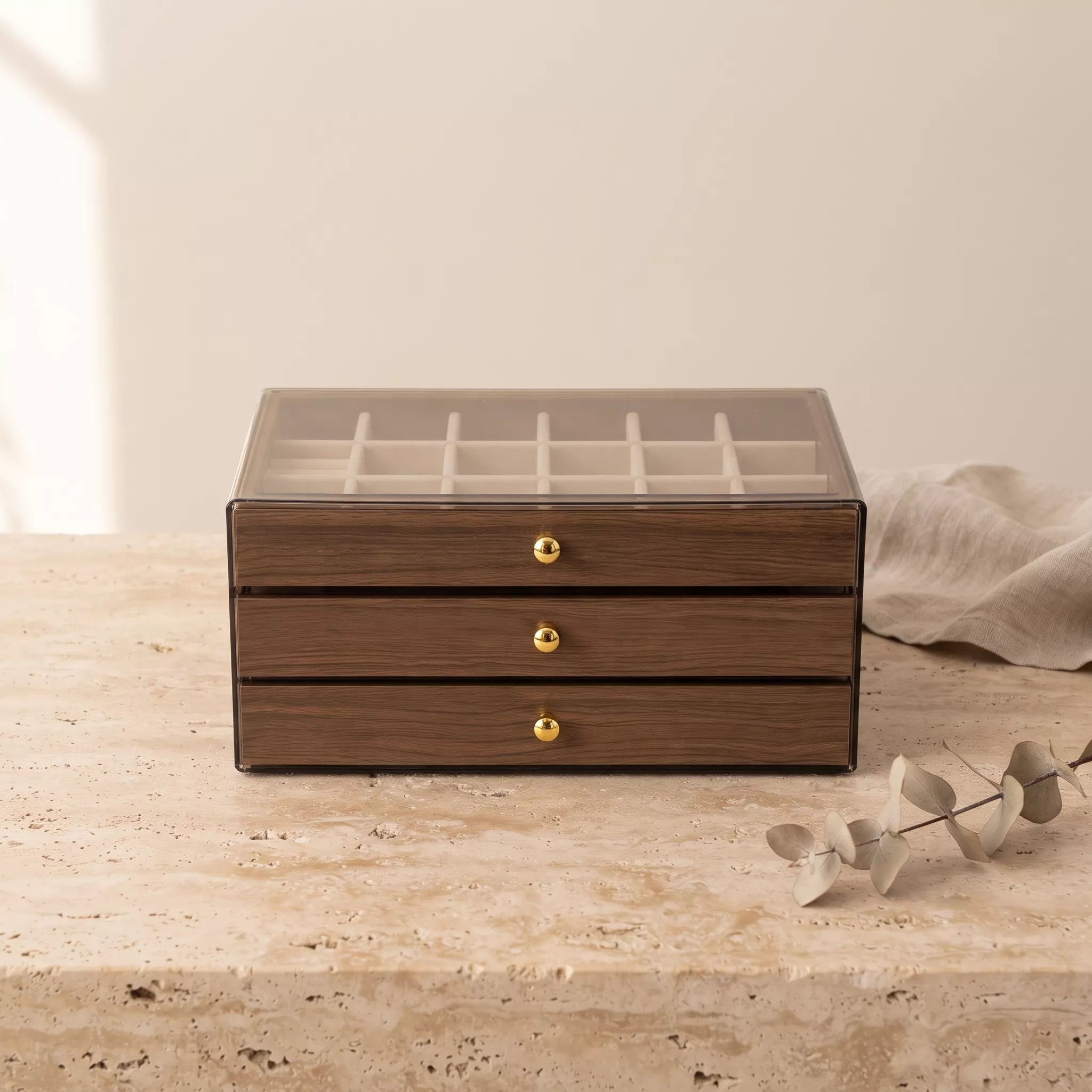 A rectangular wooden jewelry organizer box with three drawers and a clear glass lid showing an interior ring slot grid. The box has a medium brown wood grain finish with small round gold knobs and sits on a light stone surface against a warm beige background, with a draped fabric and decorative eucalyptus branch nearby.