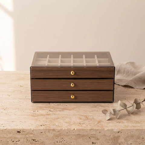 A rectangular wooden jewelry organizer box with three drawers and a clear glass lid showing an interior ring slot grid. The box has a medium brown wood grain finish with small round gold knobs and sits on a light stone surface against a warm beige background, with a draped fabric and decorative eucalyptus branch nearby.