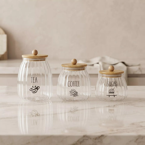 Three ribbed clear glass canisters with light wood lids and wooden knobs, labeled TEA, COFFEE, and SUGAR, arranged on a white marble countertop against a warm beige background.