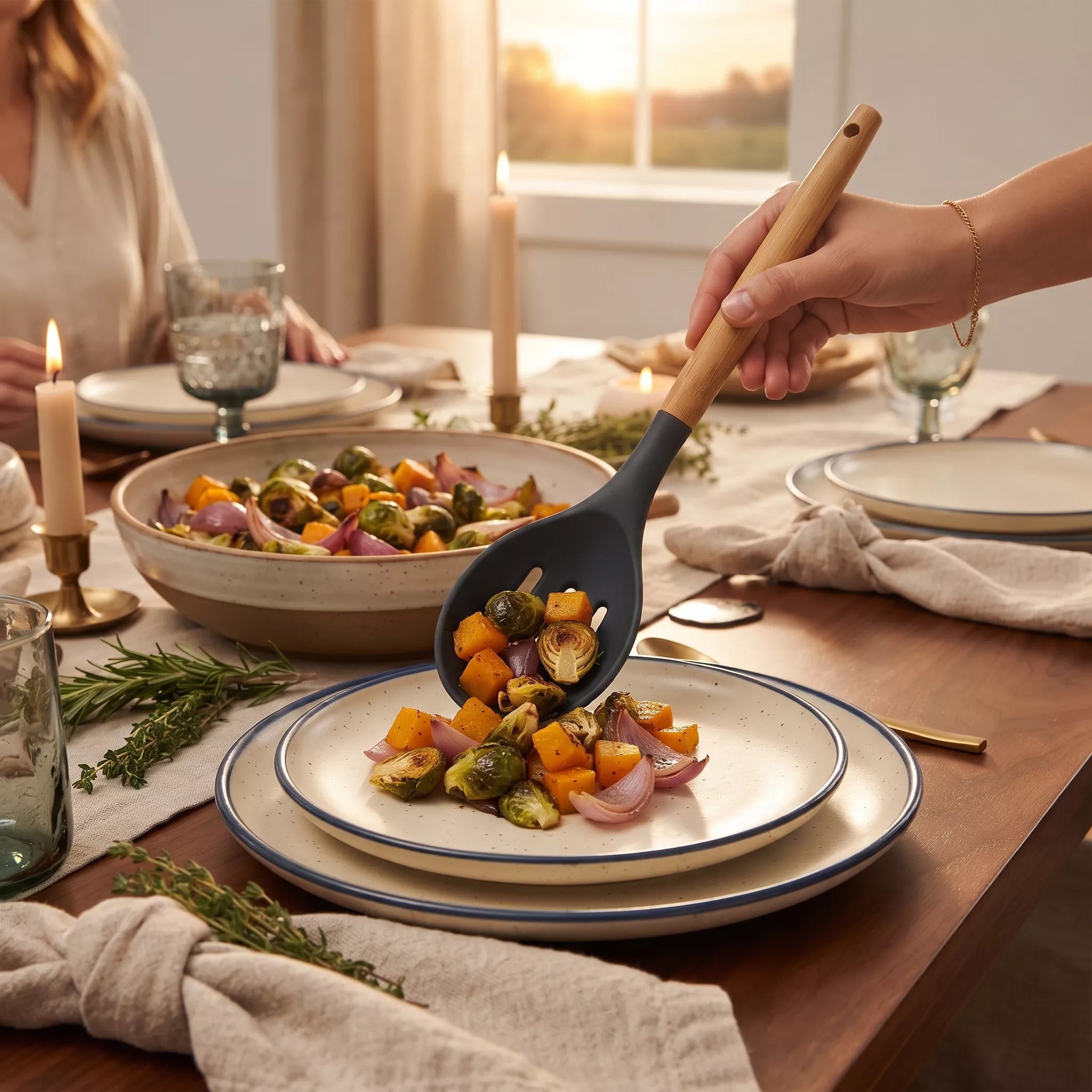 A hand uses a dark gray silicone serving spoon with a light wood handle to serve roasted vegetables from a ceramic bowl onto a stacked plate at a dining table. The scene is set in a warm, real dining room environment with other dishes, candles, and a window in the background.