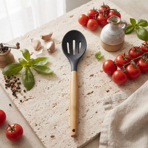 A kitchen utensil with a matte black slotted bowl and a light natural wood handle, placed on a stone board surrounded by tomatoes, basil, garlic, peppercorns, and a small ceramic pitcher.