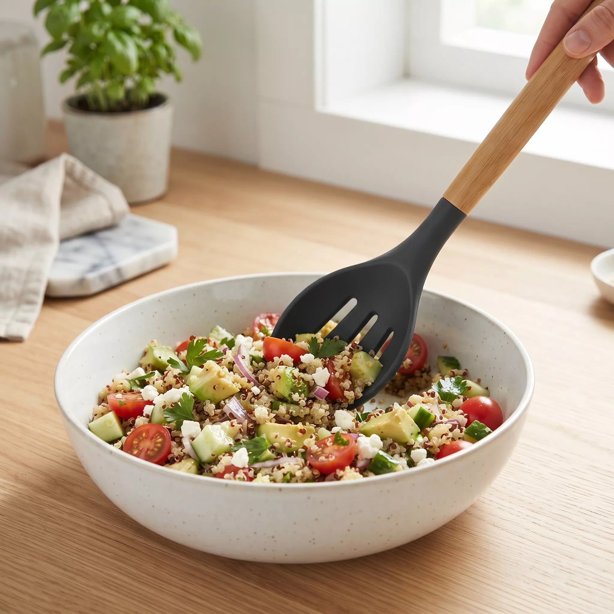 A speckled off-white ceramic salad bowl filled with a colorful quinoa salad, with a black slotted serving spoon featuring a light wood handle being lifted by a hand. The bowl sits on a light wooden table in a bright kitchen setting.