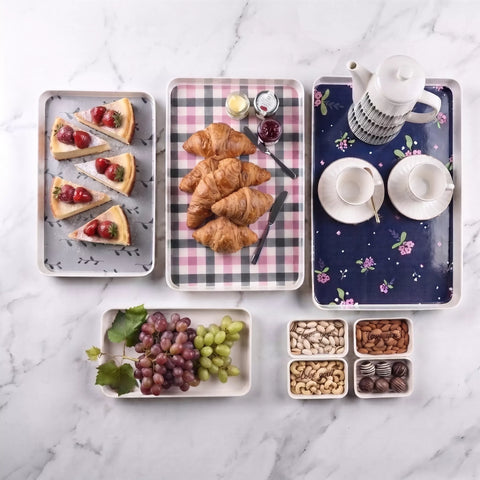 A styled flat-lay of multiple rectangular serving trays on a white marble surface, arranged with food and tableware. The trays include slices of berry tart, croissants, jam, a white teapot, two teacups, grapes, nuts, and chocolates.