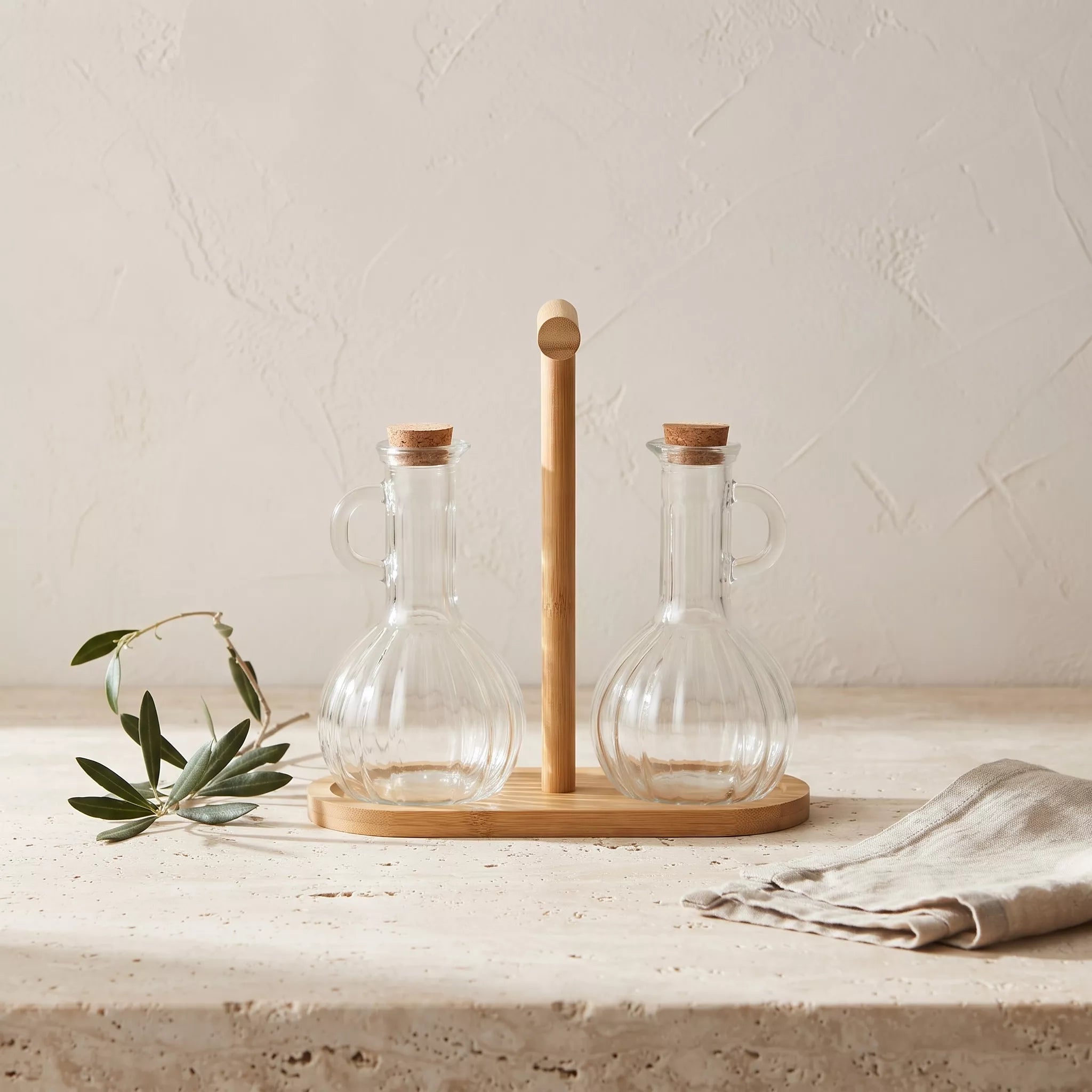 Two clear glass oil and vinegar bottles with cork stoppers and loop handles, displayed on a light wood stand with a matching wooden base, set on a stone surface with a linen cloth and olive branch.