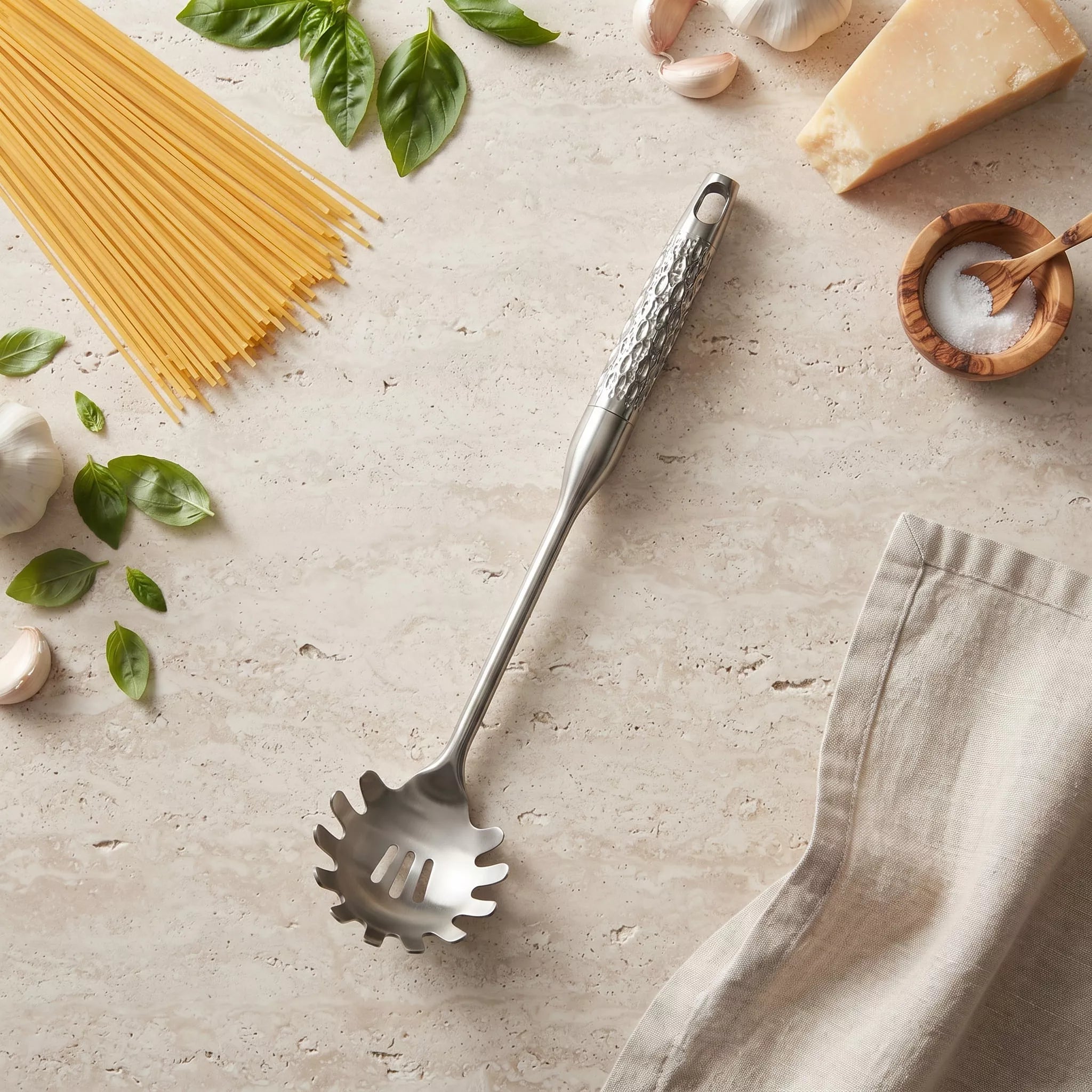 A stainless steel spaghetti server with a textured handle, styled on a light stone surface with pasta, basil, garlic, cheese, and a linen napkin.