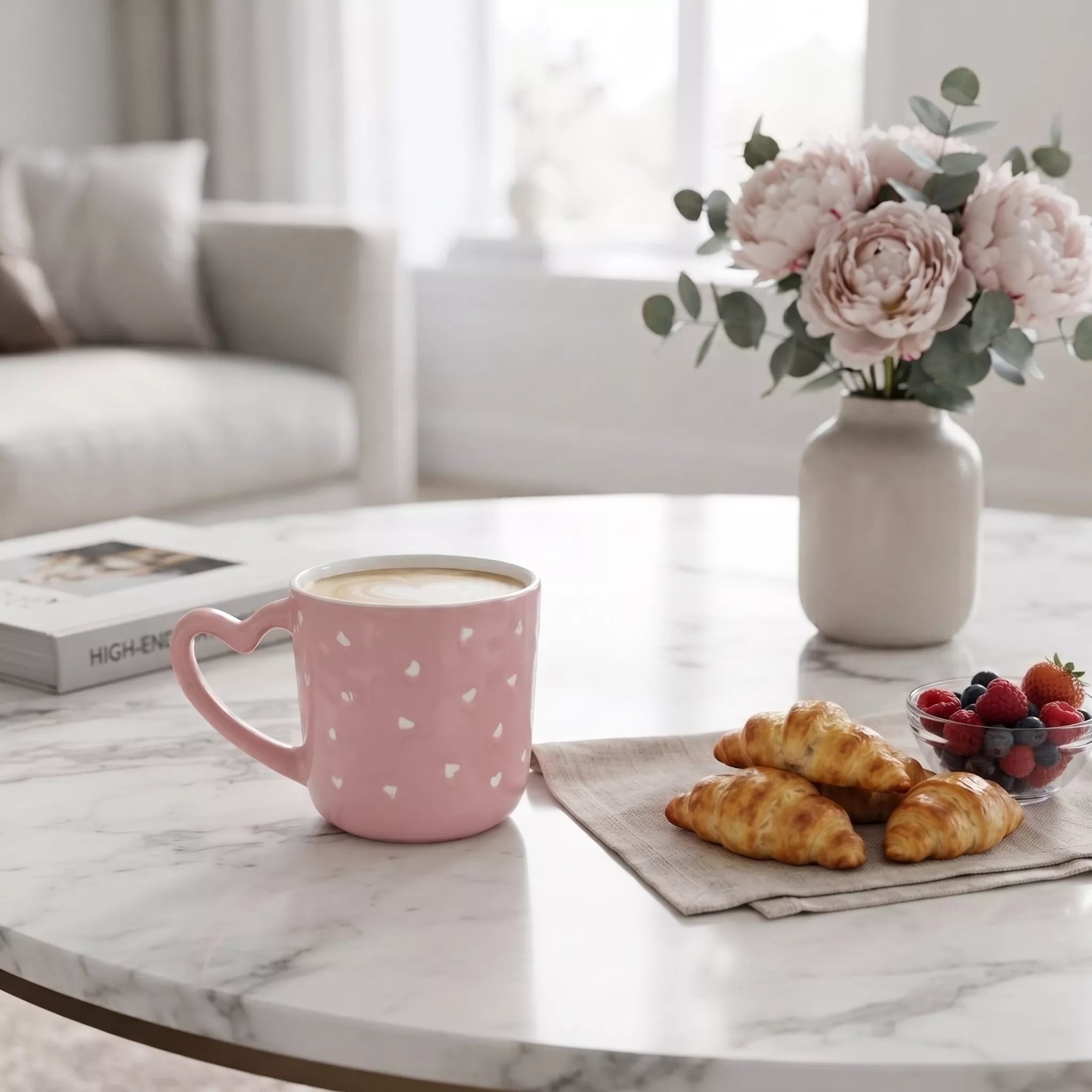 A pink ceramic mug with white heart patterns sits on a marble table in a bright, modern living room setting, styled with croissants, a bowl of berries, and a vase of pink flowers.