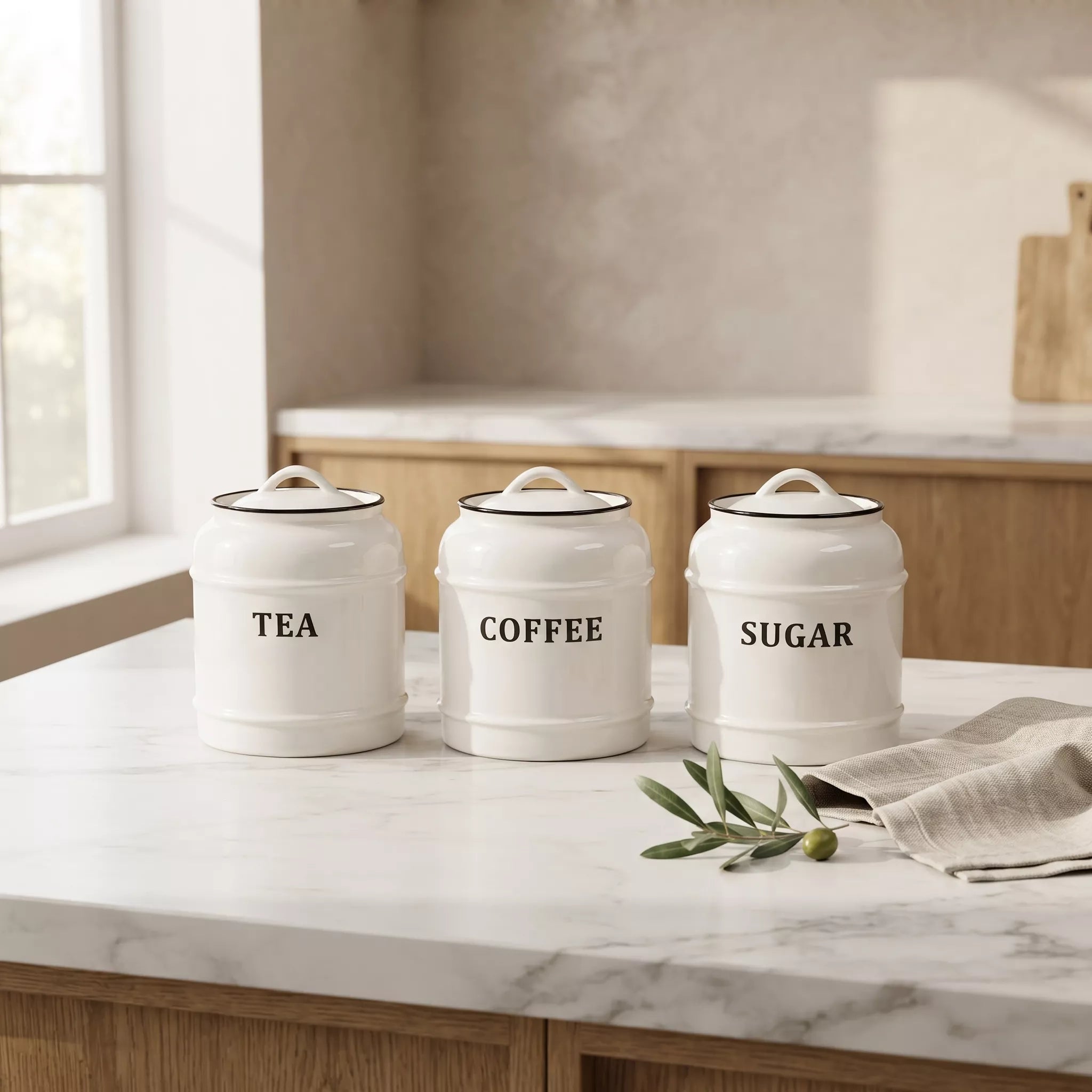 Three white ceramic canisters with lids labeled TEA, COFFEE, and SUGAR, arranged on a white marble kitchen countertop with a linen napkin and green sprigs.