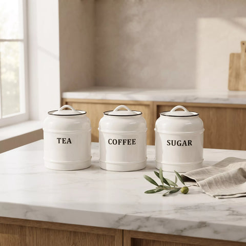 Three white ceramic canisters with lids labeled TEA, COFFEE, and SUGAR, arranged on a white marble kitchen countertop with a linen napkin and green sprigs.