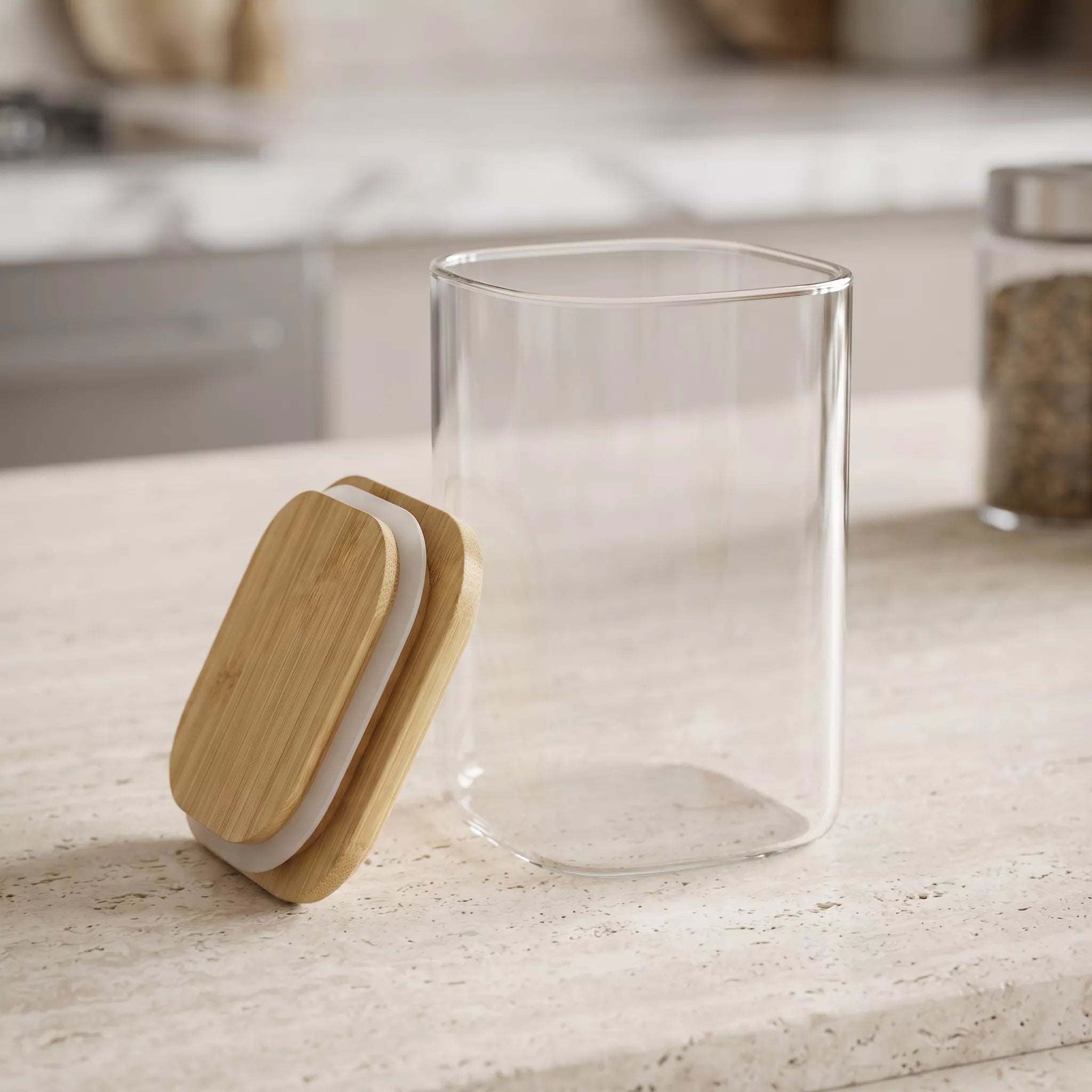Clear glass canister with a bamboo lid resting beside it on a light stone countertop in a kitchen setting.