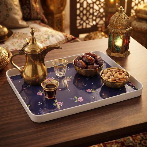 A rectangular serving tray with a white rim and a navy floral interior, styled on a wooden table with a brass teapot, two small cups, dates, nuts, and a decorative lantern in a warm indoor setting.