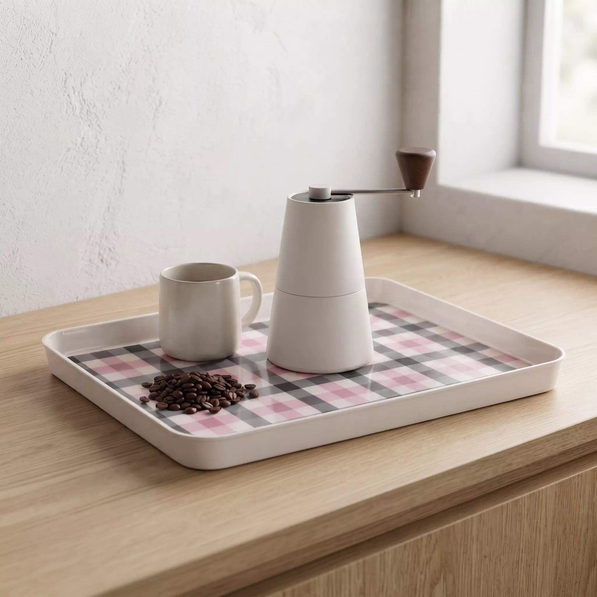 A white rectangular serving tray with a pink and gray plaid patterned bottom, styled on a light wood table near a window. The tray holds a white ceramic mug, a white manual coffee grinder, and a small pile of coffee beans.