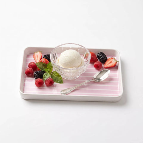 A rectangular pink tray with white stripes holds a glass bowl of vanilla ice cream, fresh berries, mint leaves, and a spoon on a clean white background.