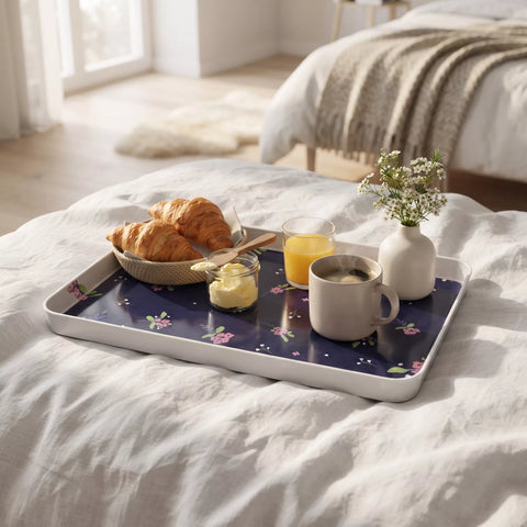 A rectangular white tray with a dark blue floral liner sits on a bed, styled with croissants, a glass of orange juice, a mug, a small jar of butter, and a white vase with baby's breath.