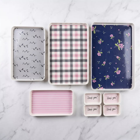 A set of rectangular storage trays and small dish containers arranged on a white marble surface. The trays feature different printed designs: one with small black bird silhouettes on light gray, one with pink and gray plaid, one with a navy floral print, one with pink horizontal stripes, and four small containers with 'Love you' text on a striped background.