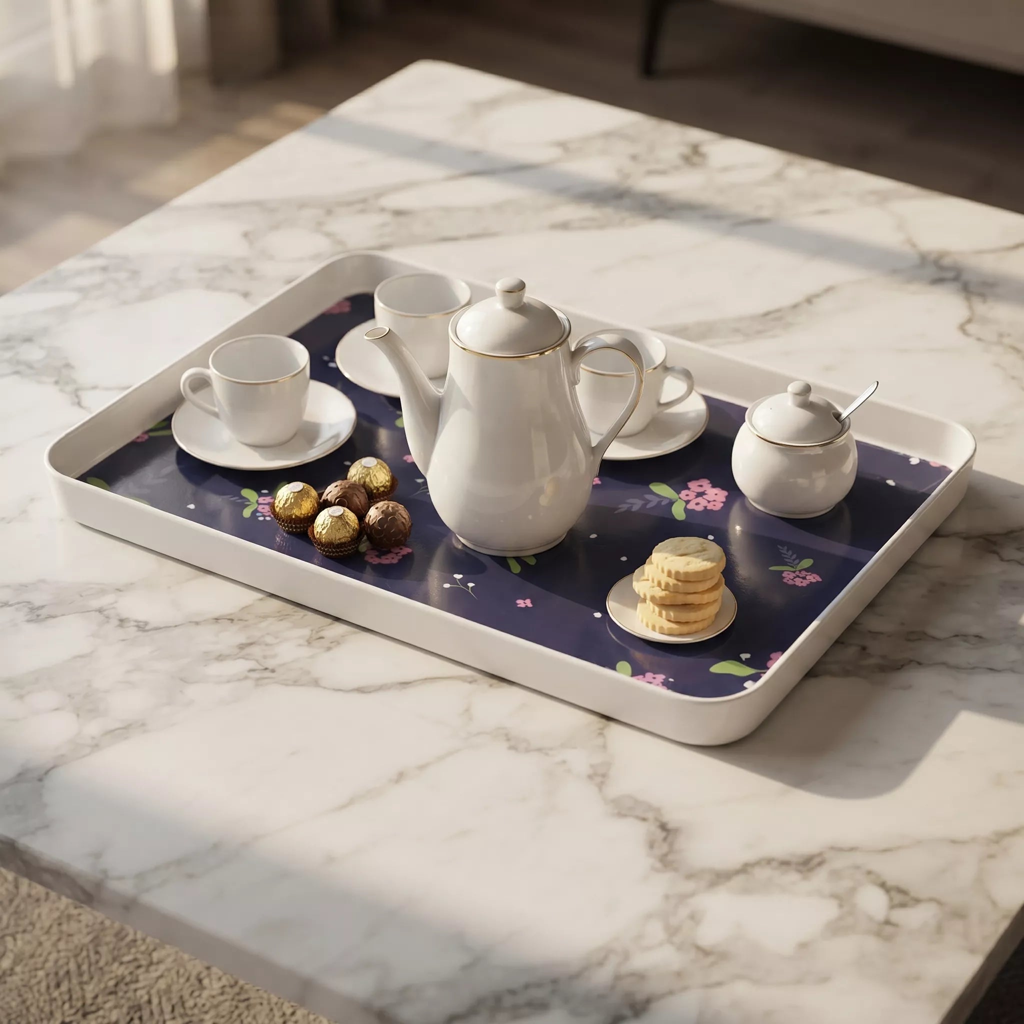 A white rectangular serving tray with a dark blue floral liner holding a white teapot, two teacups on saucers, a small creamer jar with lid and spoon, chocolates, and cookies, set on a white marble table.