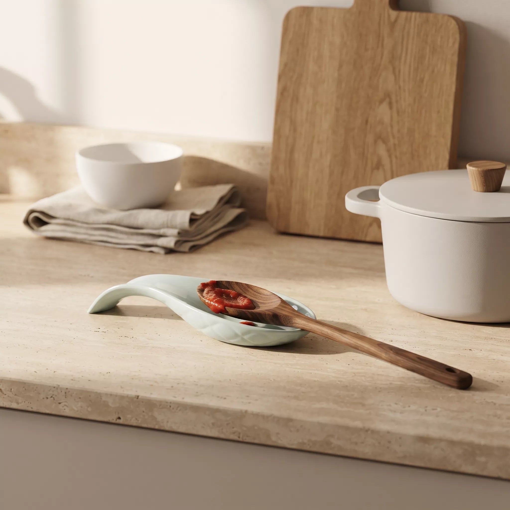 A ceramic spoon-shaped dish in a pale mint green glaze holds a wooden spoon with sliced red peppers, styled on a beige stone countertop with a white pot, bowl, and wooden cutting board in the background.