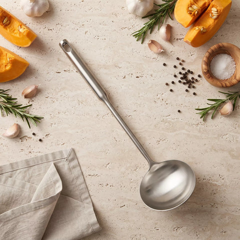 A stainless steel kitchen ladle with a long handle is arranged diagonally on a textured beige stone surface, surrounded by cooking ingredients including sliced orange pumpkin, garlic cloves, rosemary sprigs, peppercorns, and a folded beige cloth napkin.