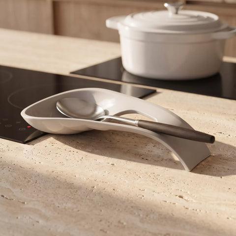 A modern curved kitchen spoon rest in a light gray ceramic finish, holding a stainless steel serving spoon with a dark handle, set on a beige travertine countertop with an induction cooktop and a white pot in the background.