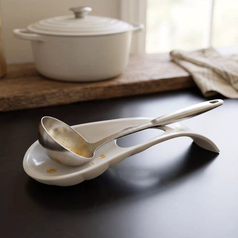 A white ceramic soup spoon rest with a stainless steel ladle resting inside it, placed on a dark tabletop with a blurred kitchen pot and cloth in the background.